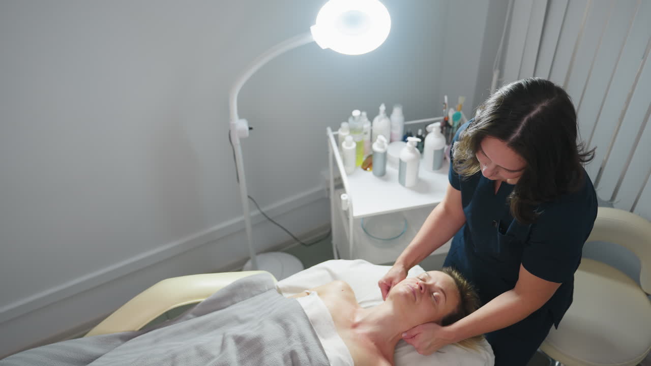 High angle view of facialist gliding fingertips along side of patient neck toward shoulder during relaxing spa session in bright clinic room with subtle soft product trolley in background
