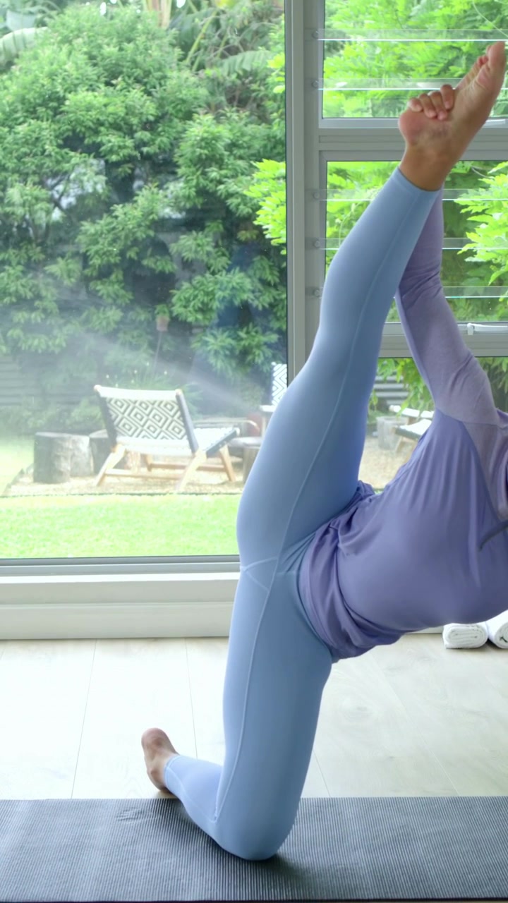 Vertical video: Pulling foot to torso kneeling on mat, woman in top balancing by window, copy space