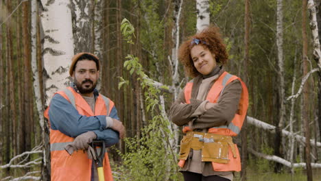 african american woman activist and arab coworker posing for the camera in the forest while she is crossing her arms and he is holding a shovel