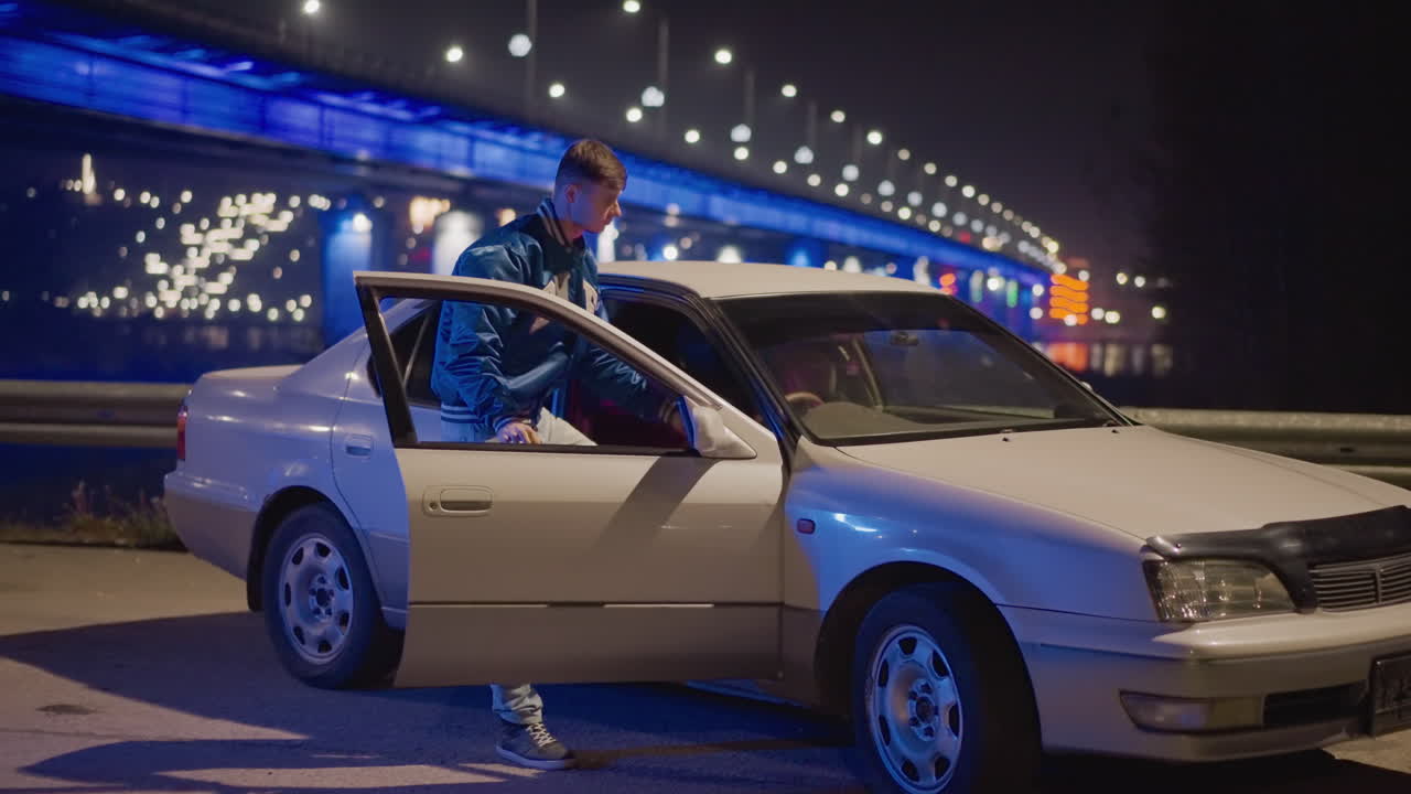 Approaching car near glowing bridge, Individual heads toward vehicle under luminous bridge, Man advances intentionally towards his car parked beside brightly lit bridge with neon reflections