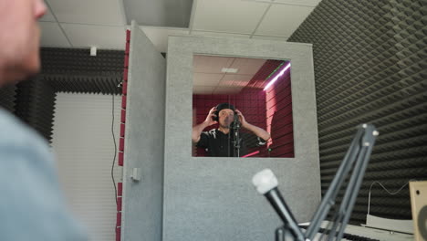 close-up of a man in a blue shirt with another man dressed in black inside a red soundproofed room. the man in black is in front of a microphone, taking off his headset indicating the end of a session