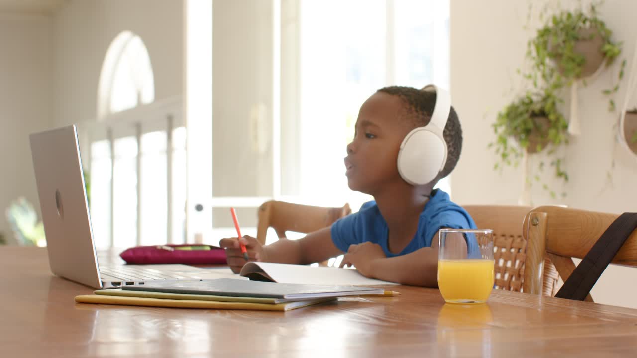 African American child at table writing in notebook, raising hand to laptop prompt, copy space
