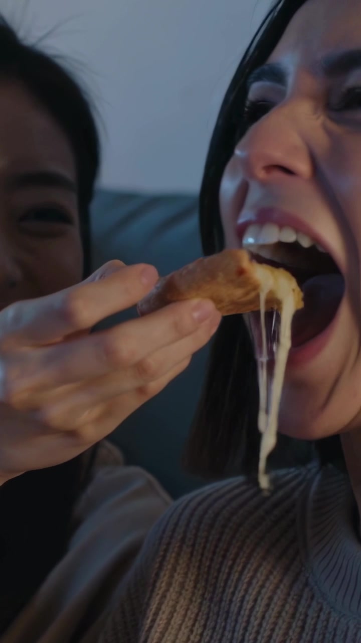 Vertical video: Guiding left woman in knits offering cheese to friend on couch for playful feeding