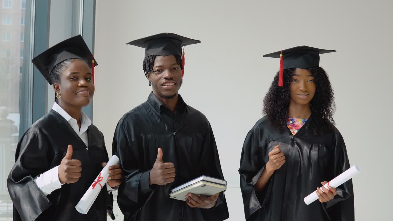 dos jóvenes mujeres afroamericanas y un joven graduado estadounidense con diplomas y libros en sus manos están de pie junto a la ventana. los estudiantes están vestidos con una túnica festiva y un sombrero de maestro