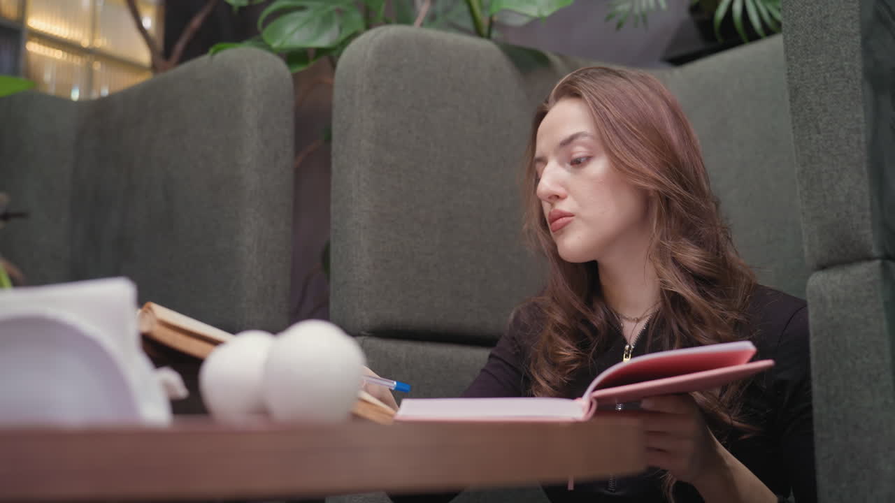 Lady in black outfit glances at open brown book then begins writing in pink notebook while seated in cozy green booth, surrounded by greenery and modern interior, maintaining calm focused expression