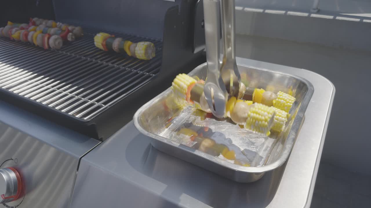 Vegetable skewers with corn, pepper, zucchini, potatoes and mushrooms are placed on a stainless steel bbq using stainless steel tongs