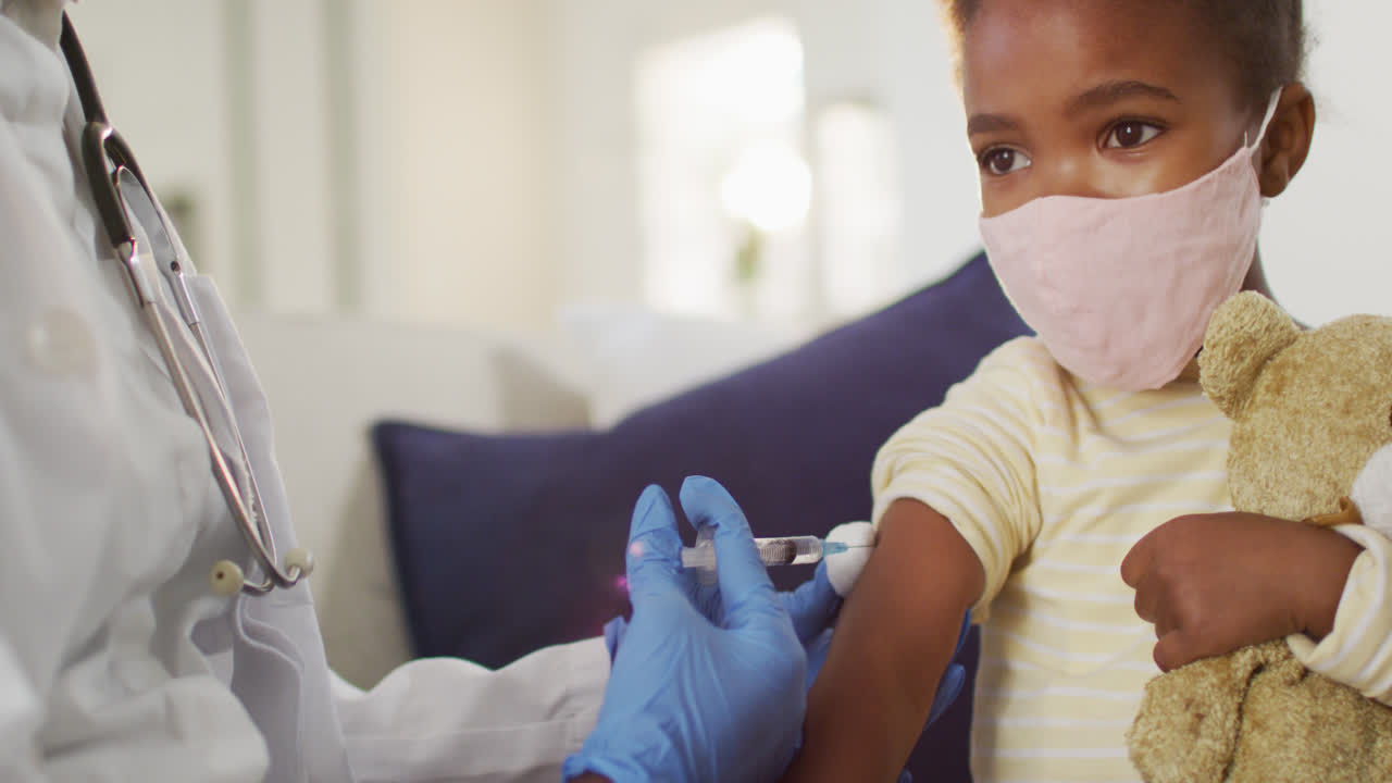 Médecin femme afro-américaine administrant la vaccination contre la COVID-19 à une fille portant un masque facial à domicile