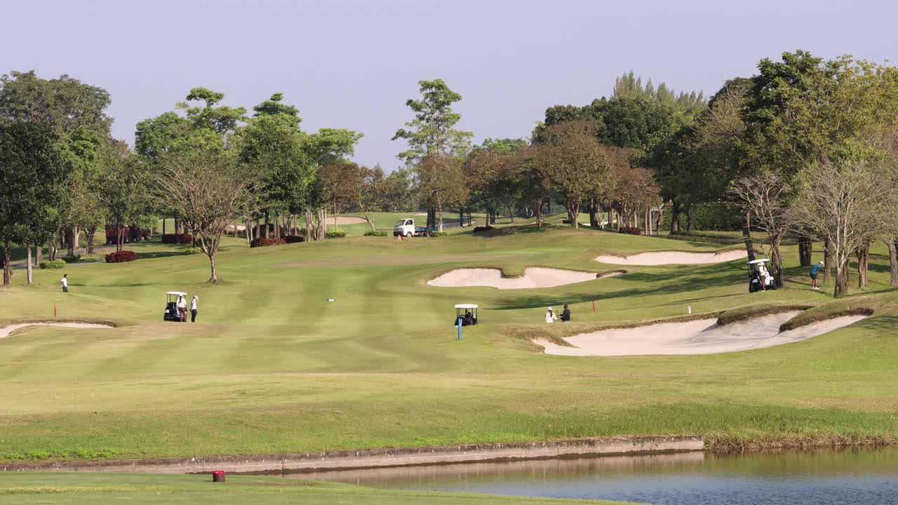 Golfers in carts traverse lush fairway with sand bunkers, water, and afternoon sunlight, wide shot