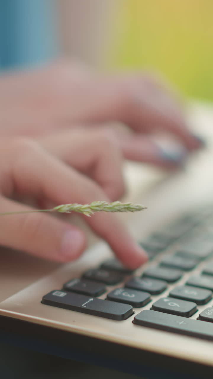 A close-up shot of a woman's hand in a blue gown, sitting on a grassy area, typing on a laptop. The image captures the moment as she gently stops a child's hand from pressing the keyboard
