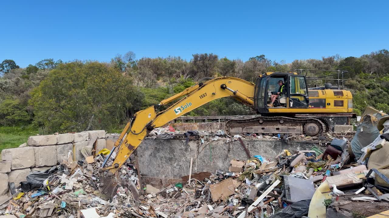 Excavator moves and lifts rubbish at landfill, bright daylight, static wide shot, natural environment