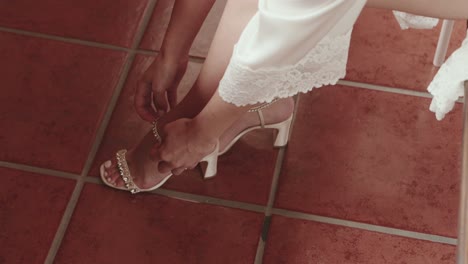 bride fastening pearl strap on elegant heels while seated in white lace robe