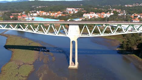 railway bridge with power lines over the ulla river with low water level, seagulls flying low, industrial buildings behind, shot of rolling overhead drone traveling forward, catoira, galicia, spain