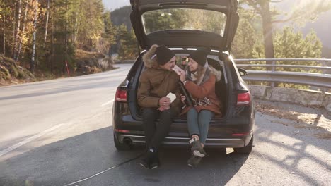 una pareja de enamorados en un viaje por carretera. el chico y la chica están sentados en el maletero abierto del coche, comiendo sándwiches. hablan, se besan. detenerse al aire libre en medio de la carretera