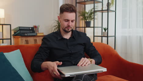 Young-bearded-man-sitting-on-sofa-closing-laptop-pc-after-finishing-business-work-in-room-at-home