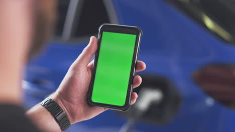 hombre cargando un coche eléctrico con un cable usando un teléfono de pantalla verde para monitorear el nivel de la batería