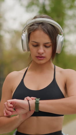 portrait view of woman wearing headphones and checking time on wristwatch outdoors in park, she is actively engaged in personal fitness routine while being mindful of her schedule
