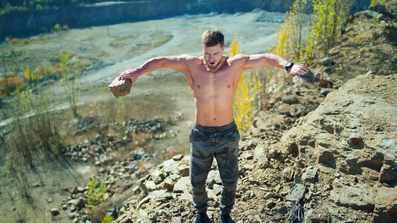 Muscular athlete doing his workout in nature. Shirtless man exercises with stones on the edge of a rocky hill in summer.