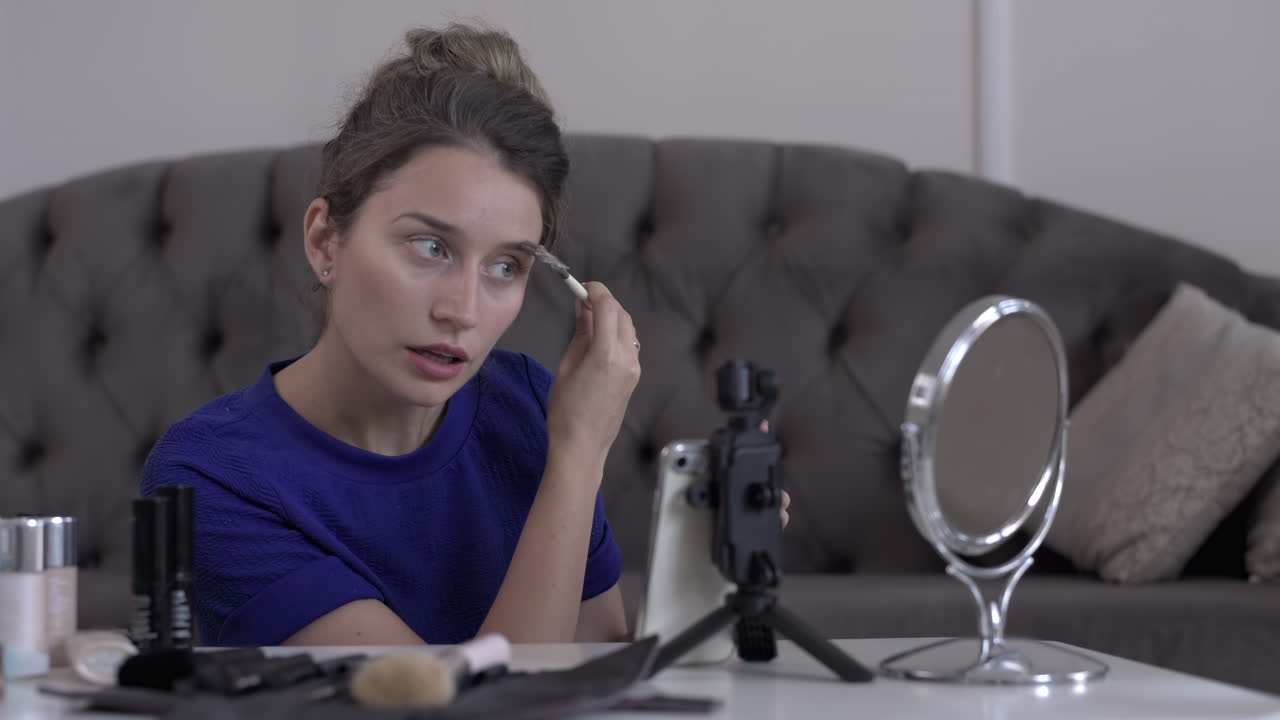 Woman in a blue T-shirt filming herself while doing a make-up tutorial at home