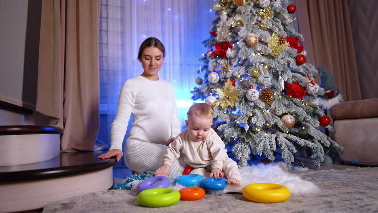 Lovely adorable toddler plays with the rings from toy pyramid. Mother sitting behind him attracts son's attention with the blue string of beads. Christmas tree at backdrop.