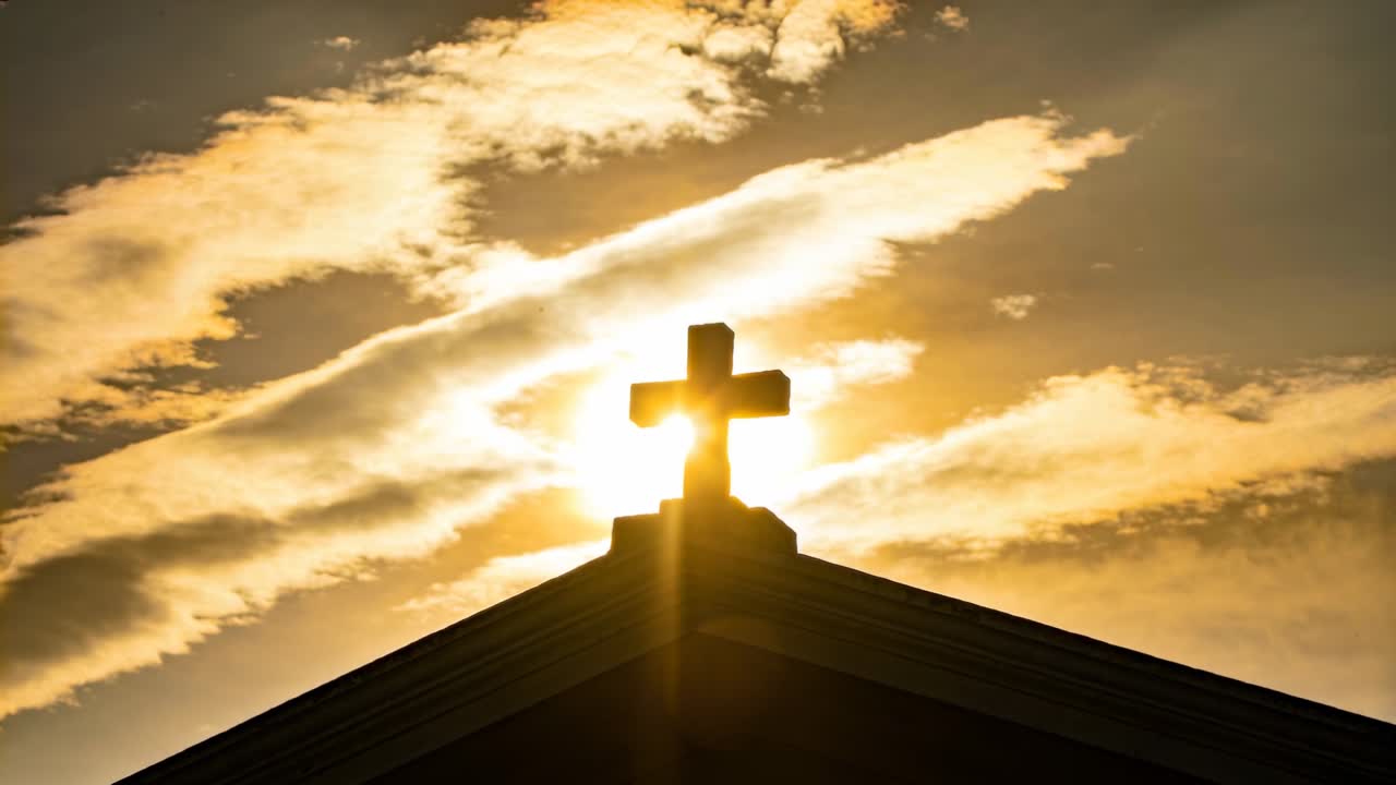Il sole cangiante che fa capolino dietro la croce di una chiesa sul tetto al tramonto, producendo un alone e un raggio verticale.