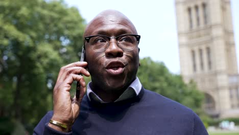 Un-Profesional-Negro-Calvo-Está-Parado-Al-Aire-Libre-Hablando-Por-Teléfono,-Usando-Gafas-Y-Una-Pulsera-Con-Una-Torre-Prominente-Y-árboles-Visibles-En-El-Fondo