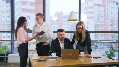 people working in the office have discussion about working moments. colleagues at table talk looking at laptop and a pair behind discuss the documents.
