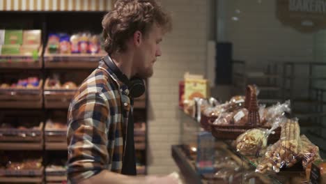 Young-guy-with-headphones-on-neck-choosing-tasty-cakes,-choosing-pastries-on-glass-showcase-in-modern-supermarket-bakery.-Side-view