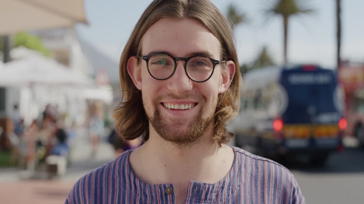 ritratto di un bel giovane geek che indossa gli occhiali che ride godendosi le vacanze estive all'aperto soleggiato che sembra calmo sulla spiaggia urbana trafficata serie di persone reali