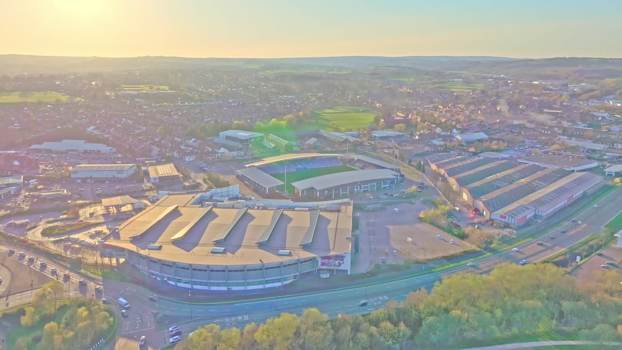 Drone shot at sunset showing the Chesterfield Football Club stadium amid warehouses, residential areas, and roads, with warm light casting a soft glow over the landscape