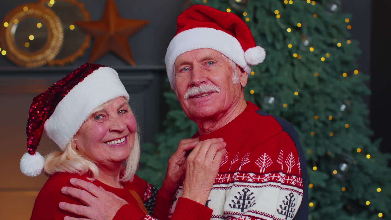 Senior couple family in Santa Claus hats celebrating Christmas looking at camera and hugging at home