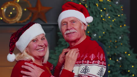 Senior-couple-family-in-Santa-Claus-hats-celebrating-Christmas-looking-at-camera-and-hugging-at-home