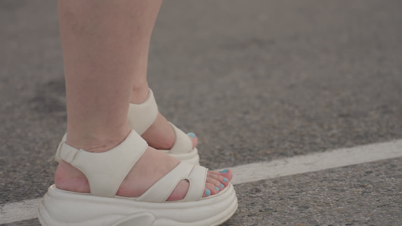 Leg view of fair skin woman walking in white platform sandals along asphalt road marked by white centre line beside green verge under soft summer light showing casual stride and gentle motion