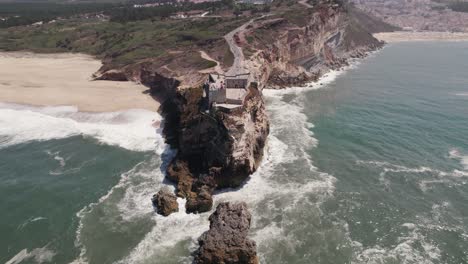 waves crashing on rocky cliffs with fortress of saint michael the archangel and nazare lighthouse suited on top