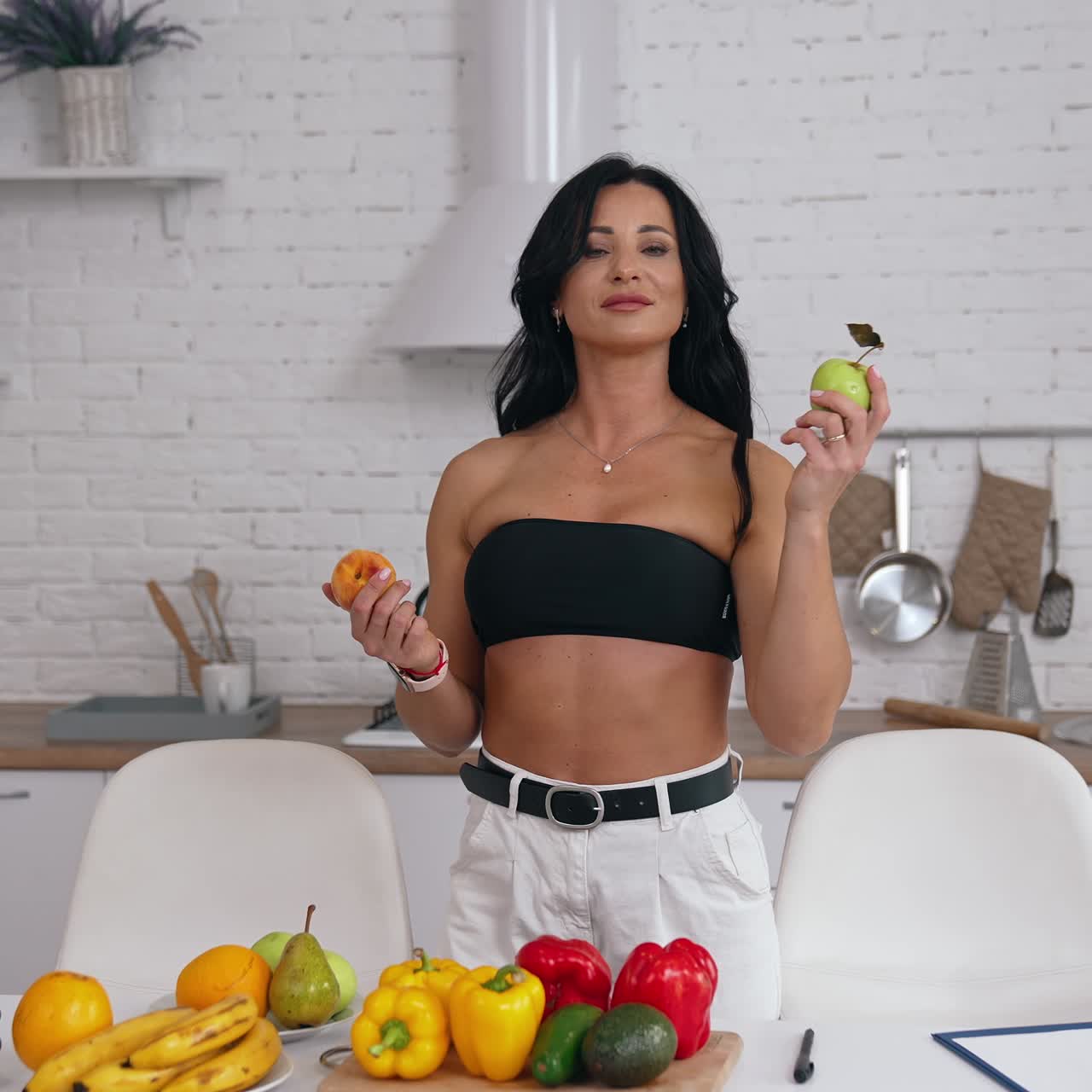 Beautiful woman in her kitchen holding fruit in both hands. Lady wearing black top and white jeans looks at fruit in her hands and then offers a peach and an apple