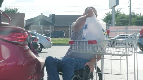 adult man with disabilities in a wheelchair puts purchases in the trunk of a car in a supermarket parking lot