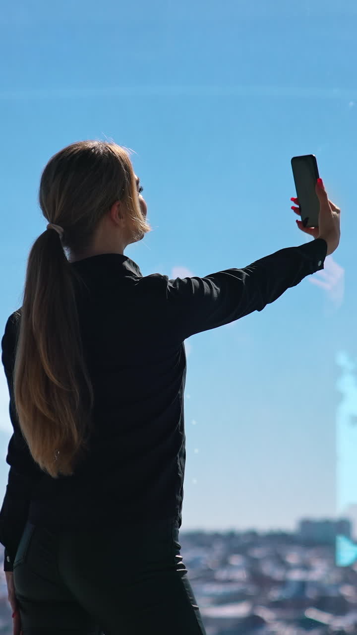 Beautiful girl posing on her phone in office. Young business woman in black doing selfie standing near large window on sky background. Back view. Vertical video