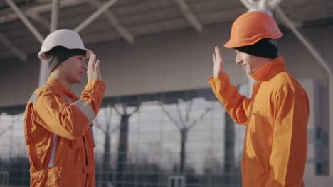 Two-construction-workers-high-five.-They-look-very-happy.-New-constructed-building-with-columns-at-the-background