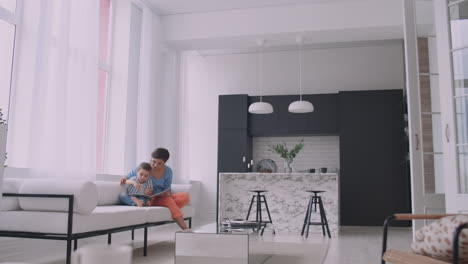 a young mother with a child reading a book sitting in a bright white interior of the house in the living room on the couch