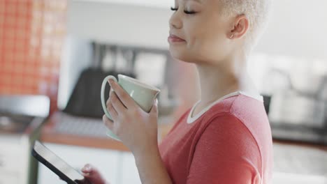 mujer biracial feliz bebiendo café y usando un teléfono inteligente en casa en cámara lenta