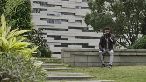 male sitting in a bench of residential district area while talking with his smartphone modern phone in outdoor green natural park