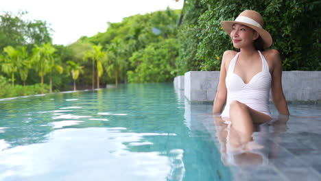 hand-held-medium-wide-shot-of-beautiful-Asian-woman-in-straw-sun-hat-and-white-swimsuit-relaxing-in-a-luxury-infinity-pool-at-a-jungle-resort