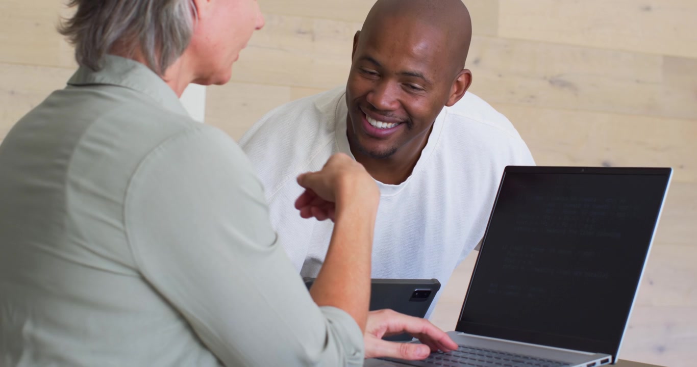 Equipe diversa trabalhando em uma mesa, mulher com relógio apontando para o laptop, usando tablet, e homem se aproximando.