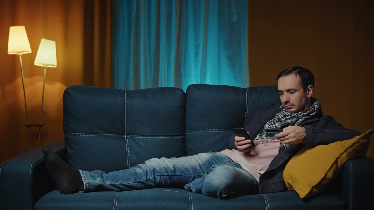 쾌적한 소파에 누워있는 남자가 신용카드로 온라인으로 지불합니다.