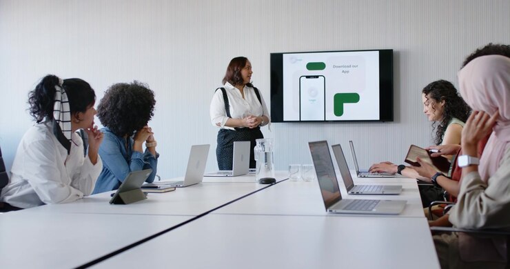 Diverse female team in meeting watching lead presenting app mockup on screen for feedback typing