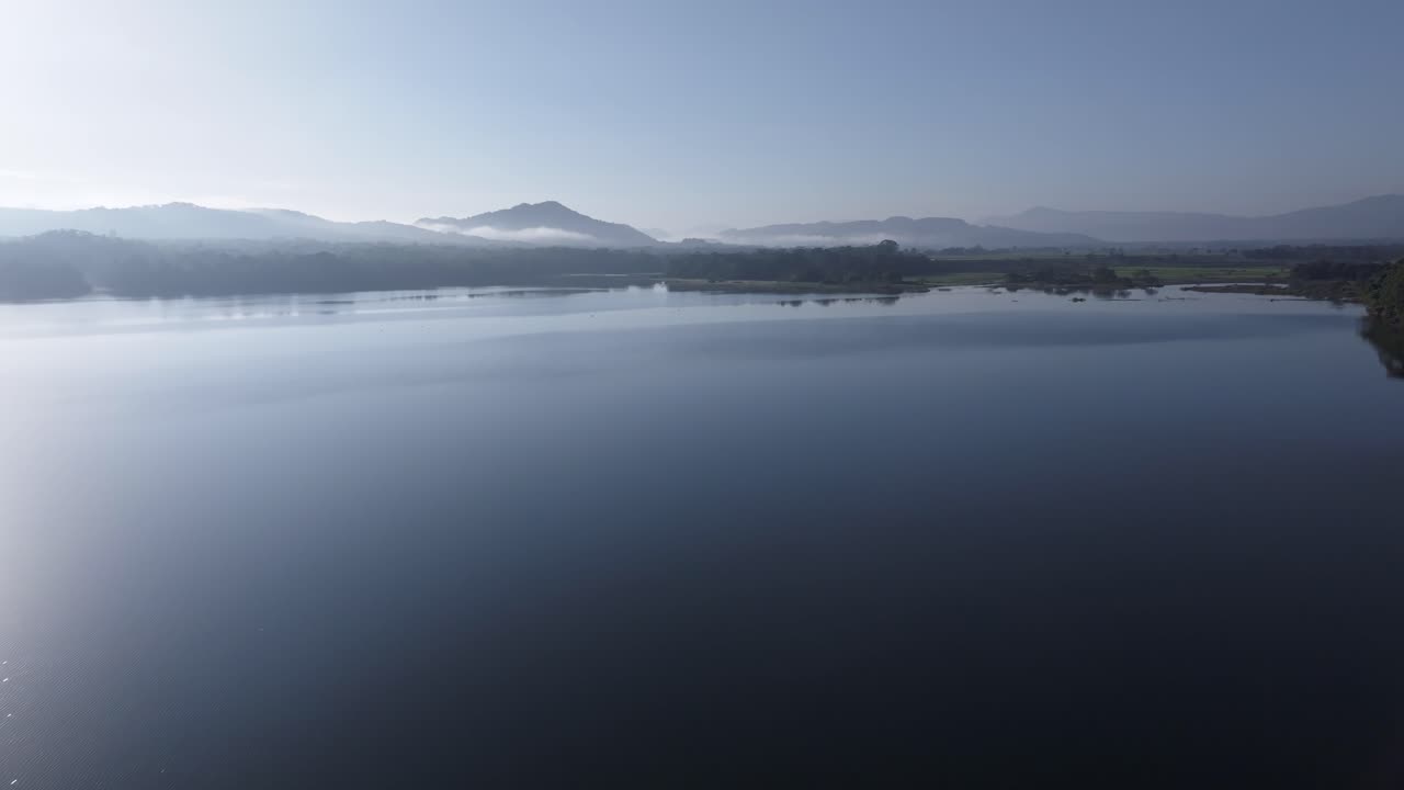 Aerial flyover sea with sun reflection during cloudy day. Mountain range and forest landscape in reservoir. Wide shot. Rincon Dam with silhouette of hills in distance.