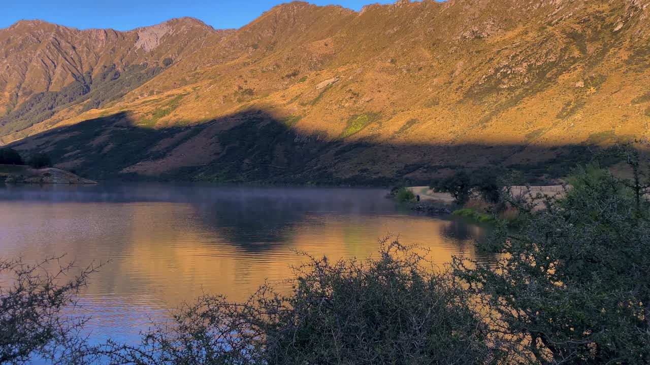 sombra de una montaña arrojada a través de un lago desde el sol de la mañana temprano, con niebla y un pescador