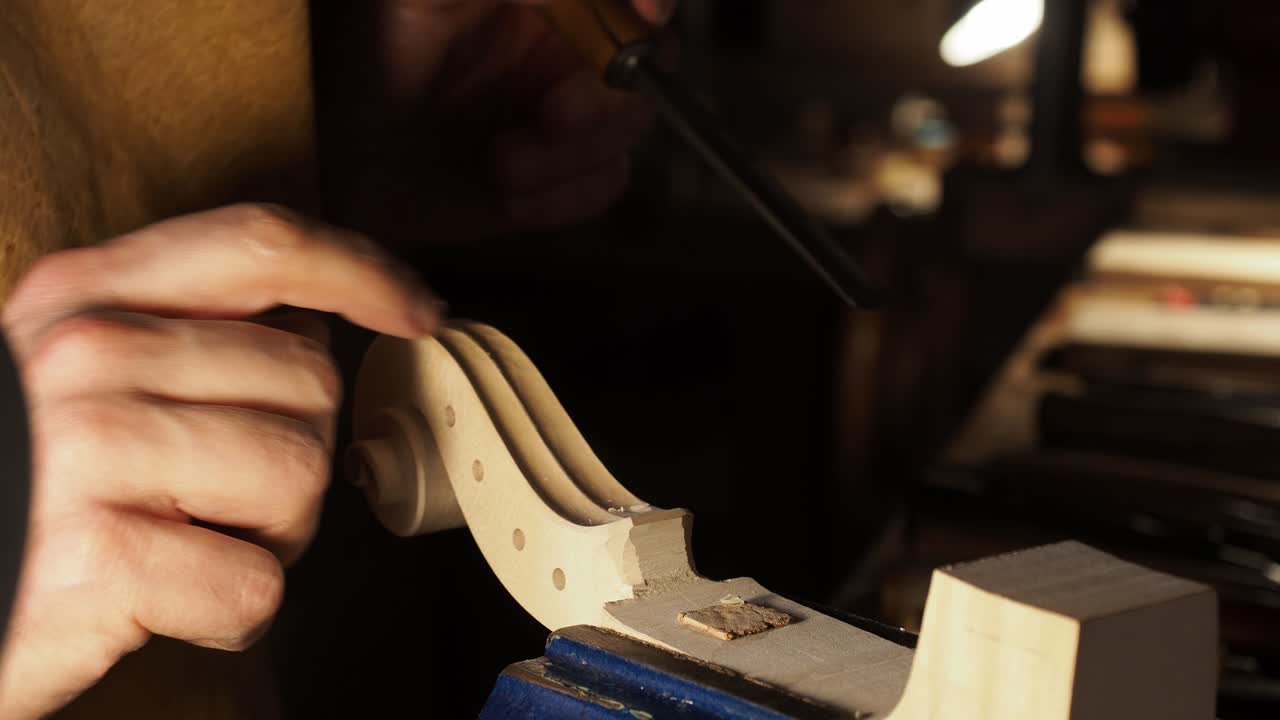 Extreme close-up of a luthier’s hands refining the scroll and peg box with a hand tool, highlighting careful wood shaping, fine detail control, and secure vice positioning