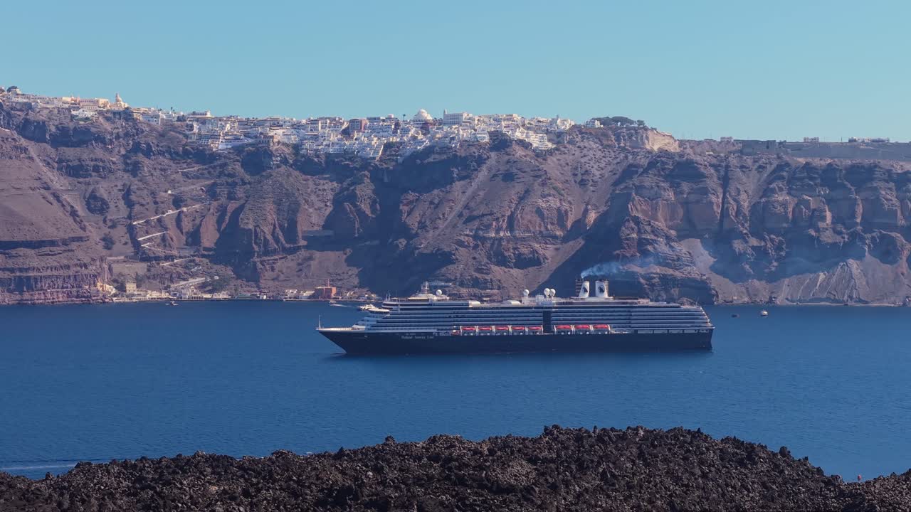 Toma de dron de un crucero navegando en la caldera de Santorini con terreno de lava accidentado en primer plano y el mar Egeo de un azul profundo.