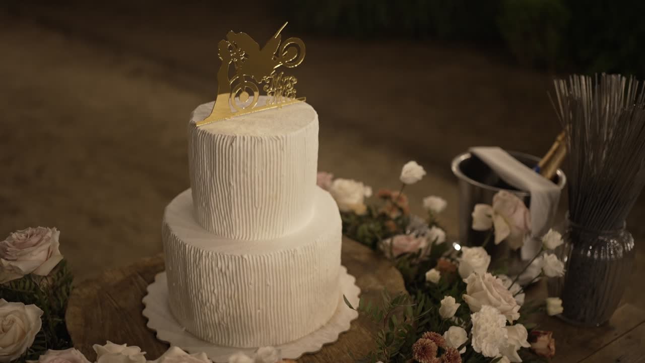 primer plano de un pastel de boda de dos niveles con decoraciones florales y un cubo de champán en una mesa rústica