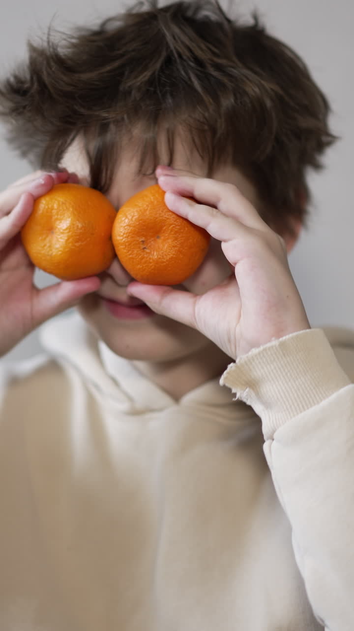Thirteen year old boy puts the tangerine in front of his eyes. Teenager showing tongue to camera. Vertical video